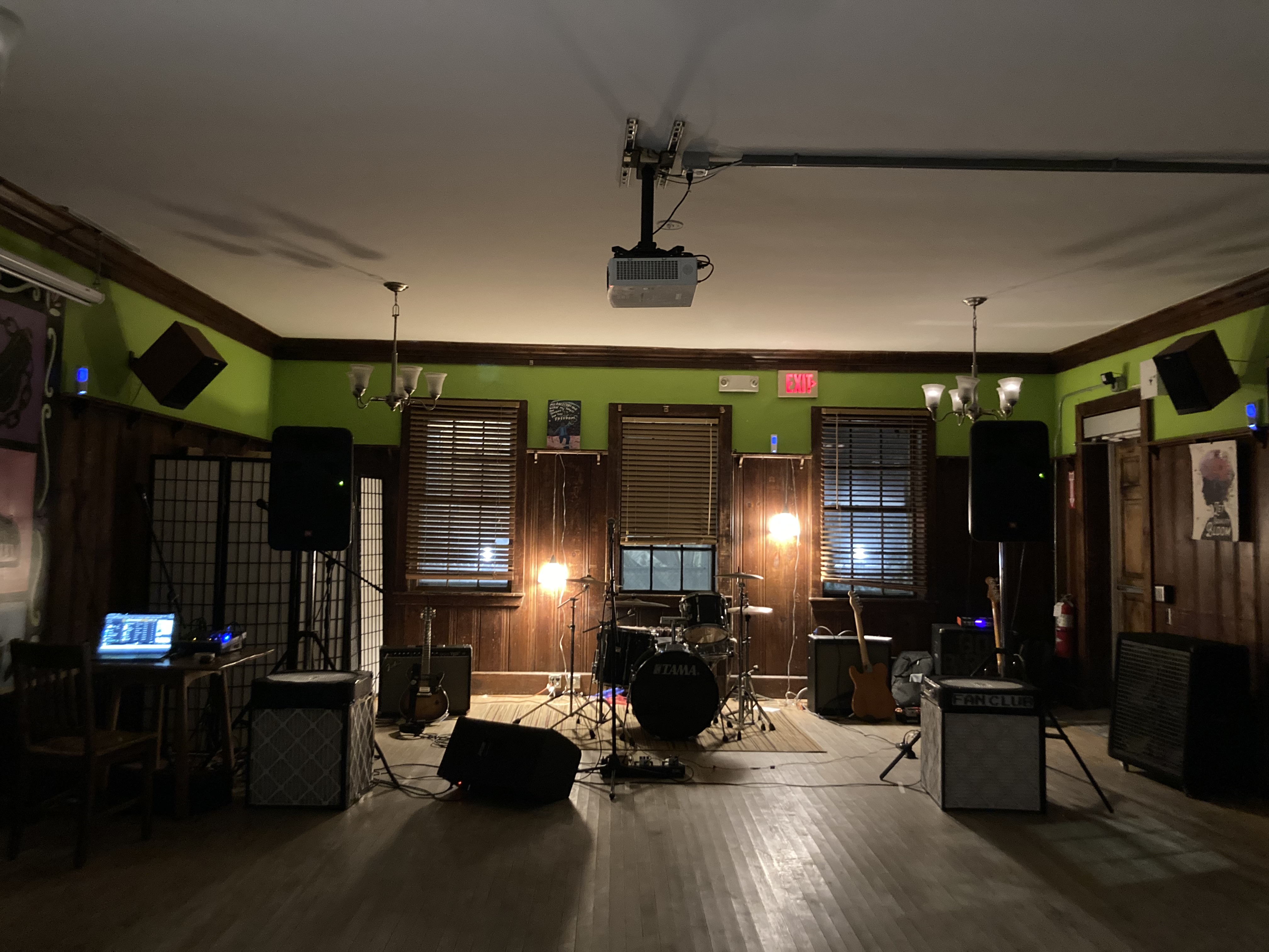 photo of a room with music equipment set up for a show including a drum kit, amps, and mic stands. there are 2 corsi-rosenthal boxes on the floor. there are also 3 small flashlight-looking items on the left, back, and right wall ledges pointed toward the viewer.